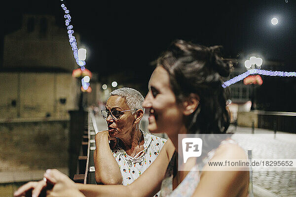 Thoughtful senior woman with friend standing by railing at night
