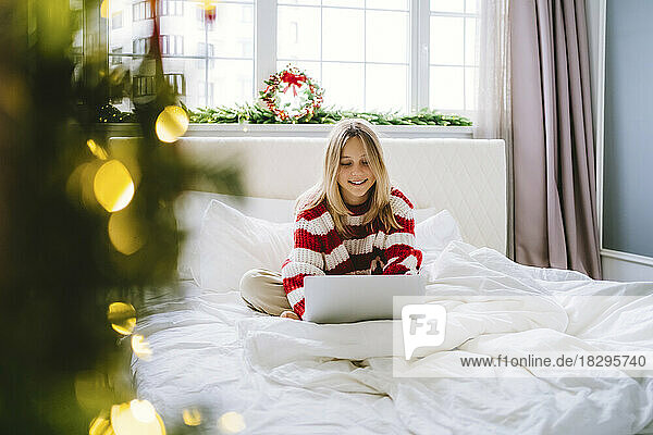 Smiling girl wearing warm clothing using laptop on bed at home