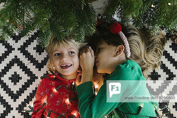 Girl whispering in sister's ear lying down with illuminated string lights near Christmas tree