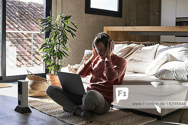 Man wearing headphones sitting with laptop on rug at home
