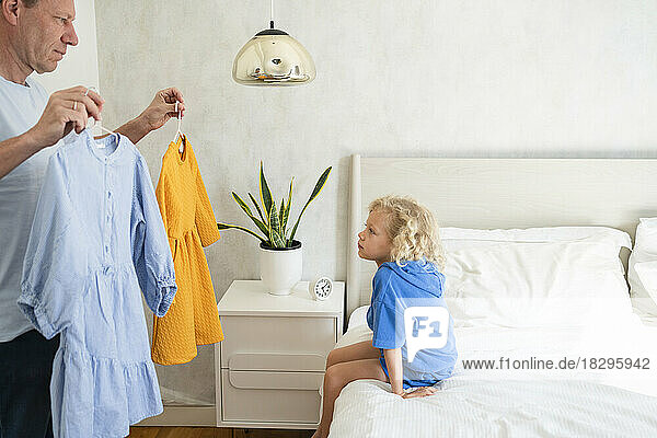 Father showing dresses to daughter sitting on bed at home