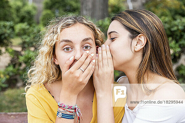 Woman whispering into shocked friend's ear in park
