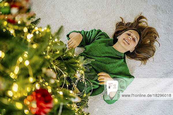 Happy girl lying on carpet under illuminated Christmas tree at home