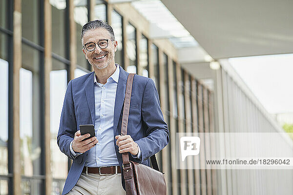 Happy mature businessman standing with smart phone outside building