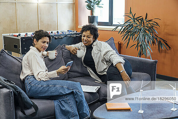 Smiling businesswoman with colleague using smart phone on sofa at office