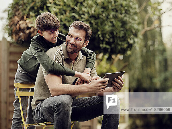 Son embraing father sitting in garden using digital tablet
