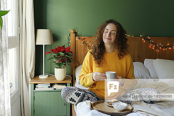 Woman with eyes closed holding cup of tea in bed at home