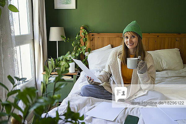 Happy woman with financial bills sitting on bed at home