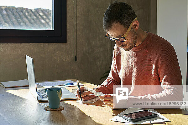 Man with pen writing and examining financial bills at desk