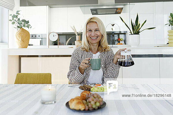 Smiling woman with coffee in cup and pot sitting at dining table