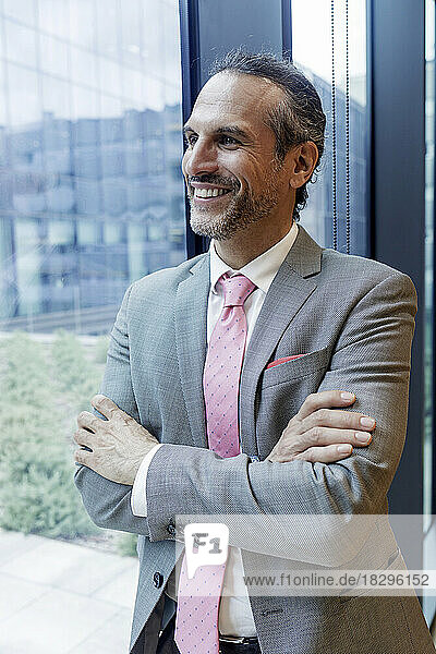 Happy businessman standing with arms crossed looking through window