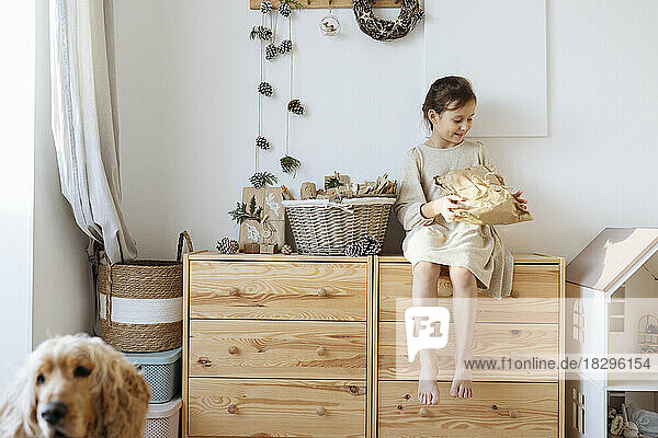 Lächelndes Mädchen hält ein Geschenk in der Hand und sitzt zu Hause auf dem Schrank