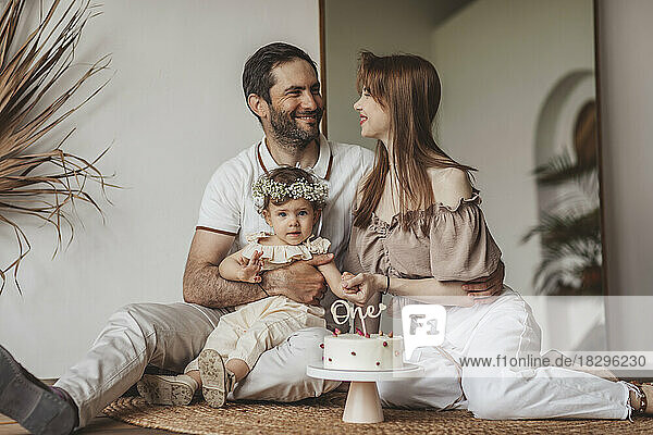 Happy parents with cute baby girl sitting in front of cake at home