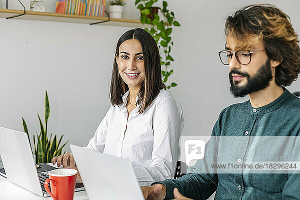 Smiling young businesswoman by colleague using laptop at desk