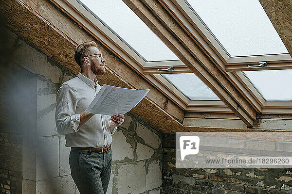 Thoughtful architect standing with blueprint looking through window at construction site