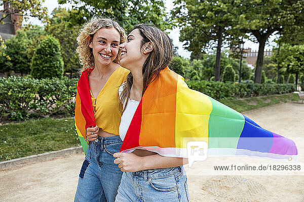 Smiling lesbian young couple covered with rainbow flag walking in park
