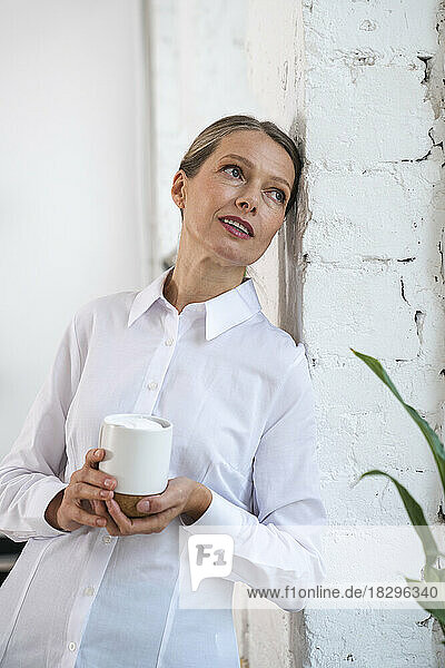 Contemplative mature businesswoman with coffee cup leaning by wall