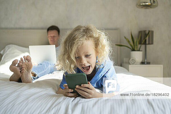 Happy girl using smart phone and father with laptop in background on bed
