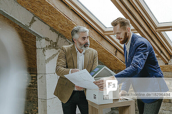 Architect explaining colleague over blueprint standing by house model at construction site