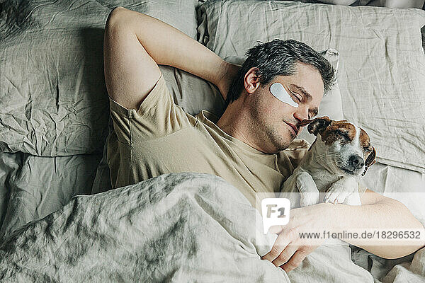 Man with under eye patches sleeping with dog on bed