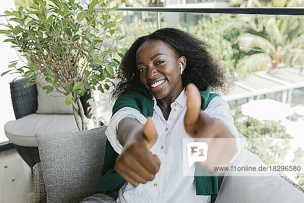 Happy woman showing thumbs up gesture sitting on sofa on balcony
