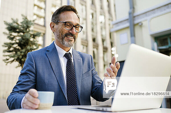 Smiling mature businessman with coffee cup gesturing on video call over laptop in cafe