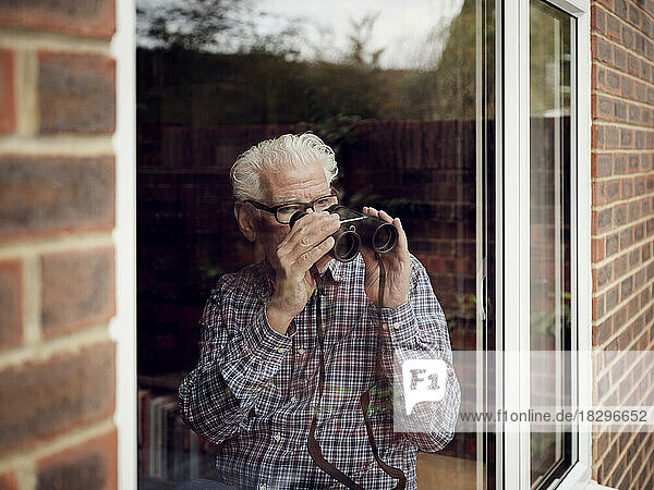 Älterer Mann schaut zu Hause mit Fernglas aus dem Fenster