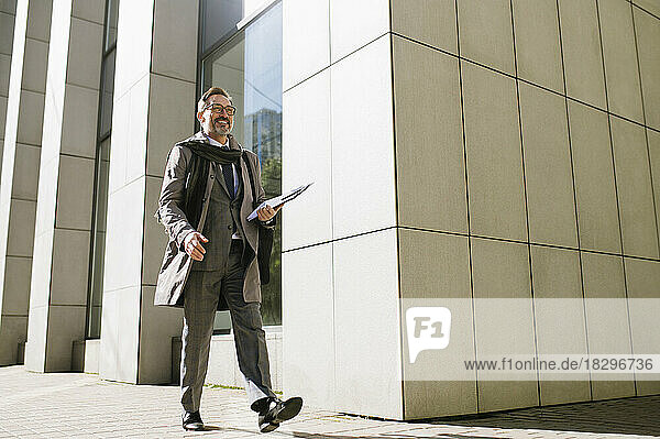 Smiling businessman with clipboard walking on footpath outside office building