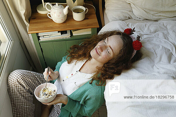 Smiling woman with eyes closed holding cereal bowl by bed at home