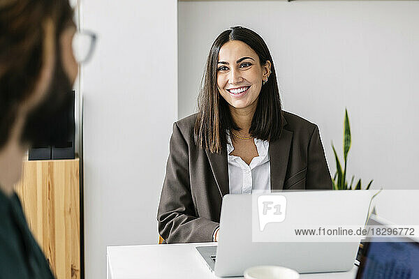 Happy businesswoman with laptop looking at colleague in office