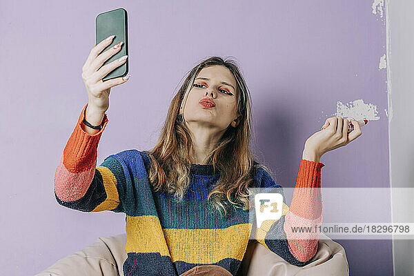 Girl puckering and taking selfie through smart phone against purple wall