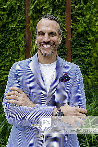 Happy mature businessman with arms crossed in front of hedge