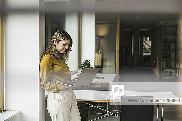 Smiling businesswoman leaning on wall and working on laptop in office