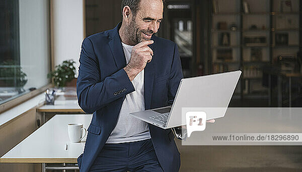 Smiling mature businessman with hand on chin and using laptop in office