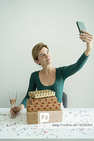 Smiling woman taking selfie through smart phone with stack of gifts on table