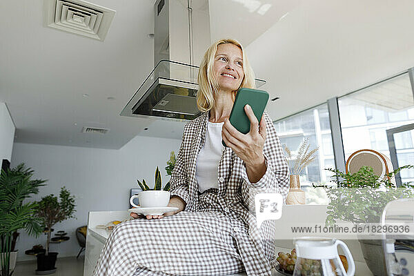 Lächelnde Frau mit Smartphone und Kaffeetasse sitzt auf der Küchentheke