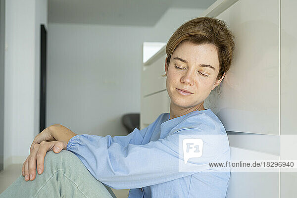 Woman with eyes closed resting in front of cabinets in kitchen