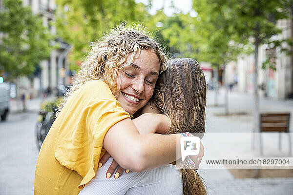 Smiling woman with eyes closed embracing friend on footpath
