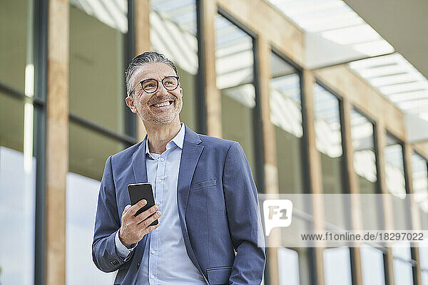 Cheerful mature businessman with mobile phone standing outside building