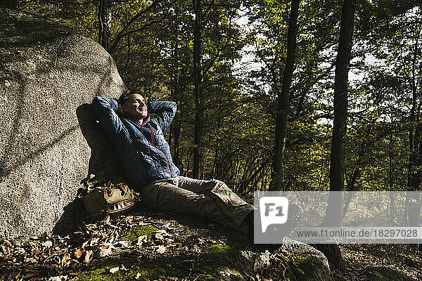 Mature man with hands behind head relaxing by rock in forest