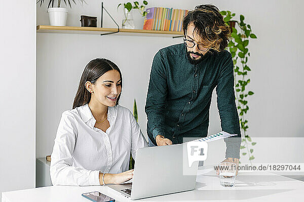 Businessman holding document discussing with businesswoman using laptop at desk