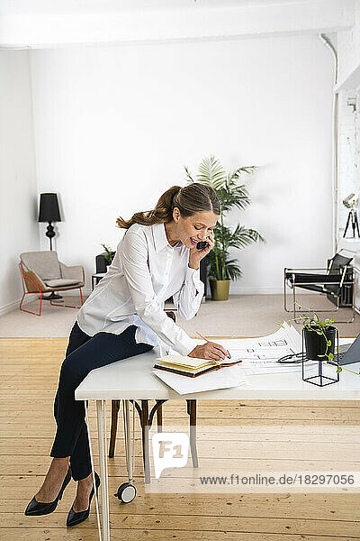 Lächelnde Geschäftsfrau  die am Schreibtisch im Büro sitzt und mit dem Mobiltelefon telefoniert und Notizen macht