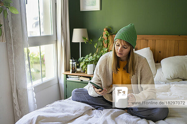 Woman using smart phone sitting with drink on bed at home