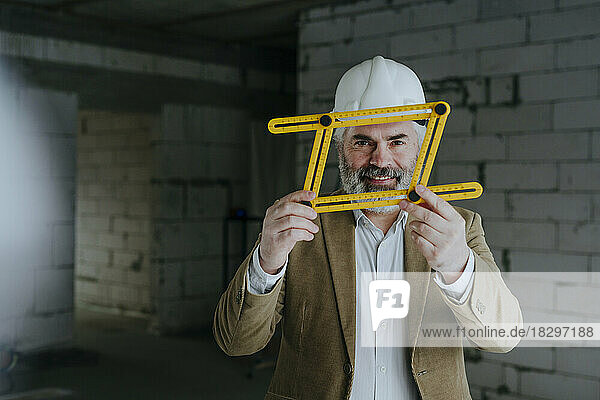 Lächelnder Architekt mit Schutzhelm  der durch ein vierfach verstellbares Lineal blickt