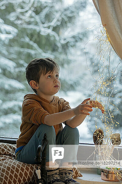 Junge berührt Lichterketten  die zu Hause auf der Fensterbank sitzen