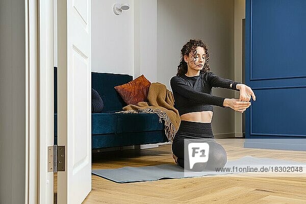 Junge Frau dehnt ihre Armmuskeln auf einer Yogamatte sitzend zu Hause