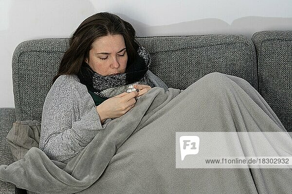 Kranke Frau  die mit einer Decke zugedeckt auf dem Sofa in ihrer Wohnung Fiebertabletten einnimmt