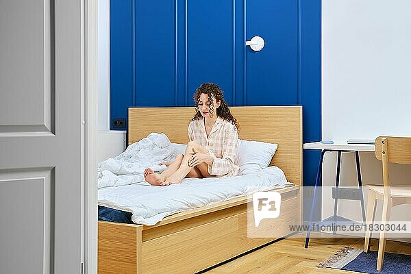 Junge Frau trägt beruhigende Aftershave-Creme auf ihre Füße auf  während sie im Bett im Bademantel sitzt