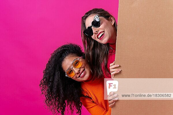 Zwei lächelnde schöne Freundinnen halten ein Pappschild auf einem rosa Hintergrund