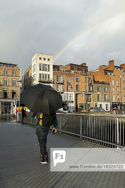 A man crosses a bridge in Dublin underneath an umbrella as a rainbow appears in the sky. Dublin  Ireland  Europe